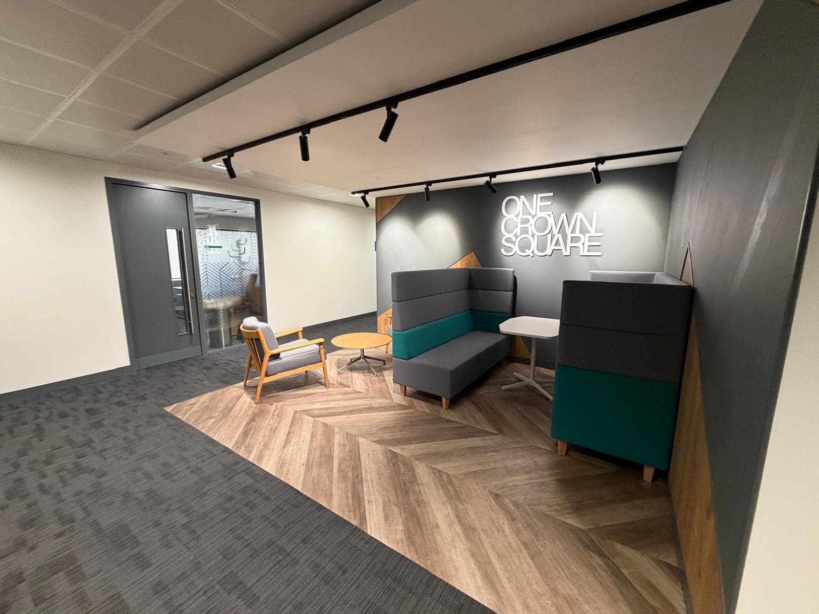 High-backed teal and grey booth seating on a herringbone timber floor, below an illuminated One Crown Square sign on a charcoal wall