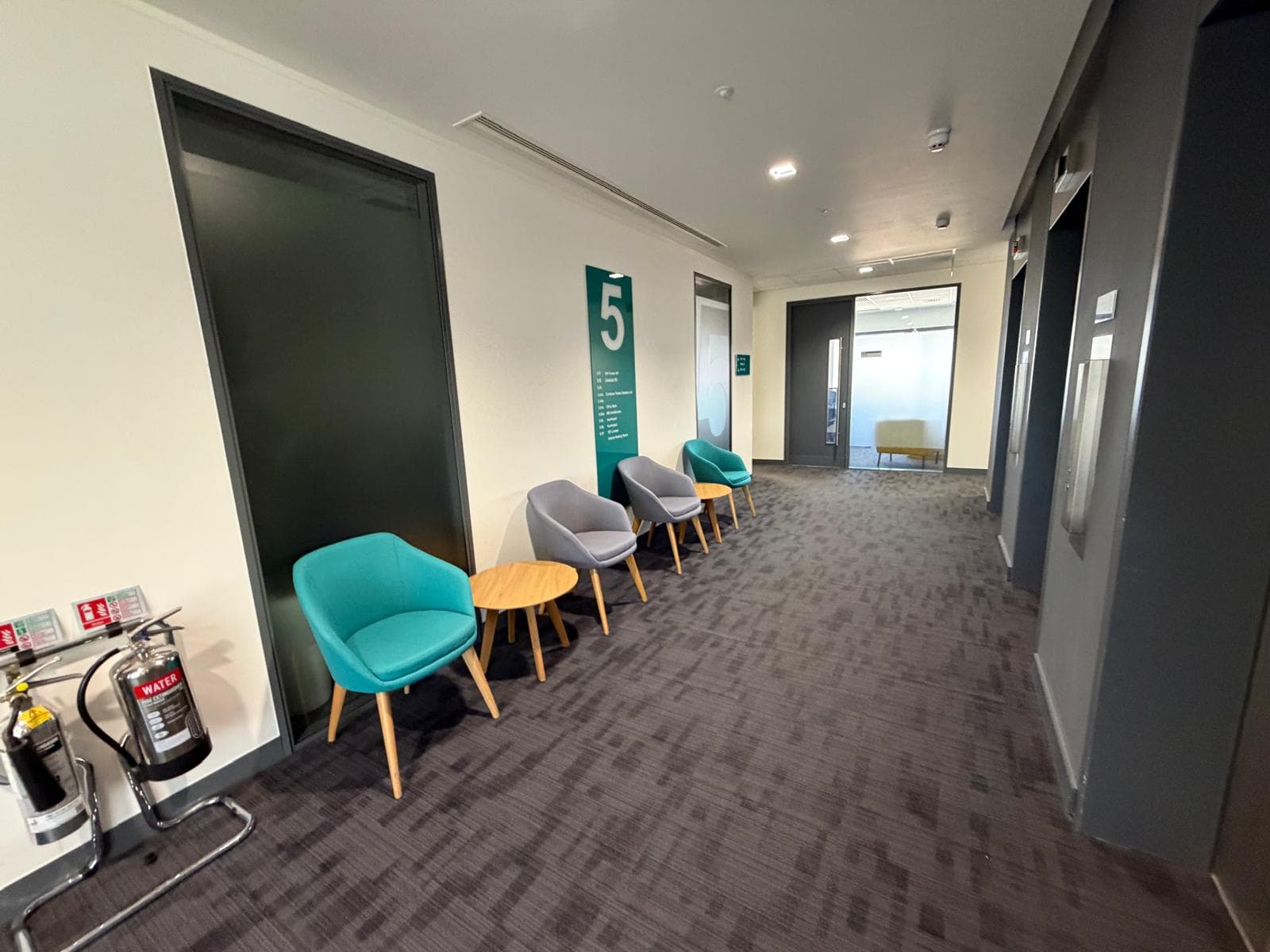 Fifth floor lobby seating, with teal and grey armchairs and a round timber table beside the floor-number column