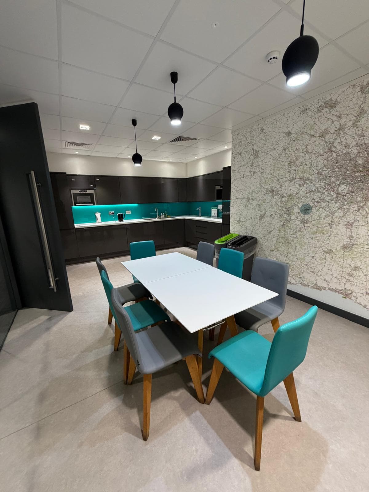 A shared kitchen and dining space with a white table and teal chairs, dark units with a lit teal splashback, and a printed map covering the full height of the end wall