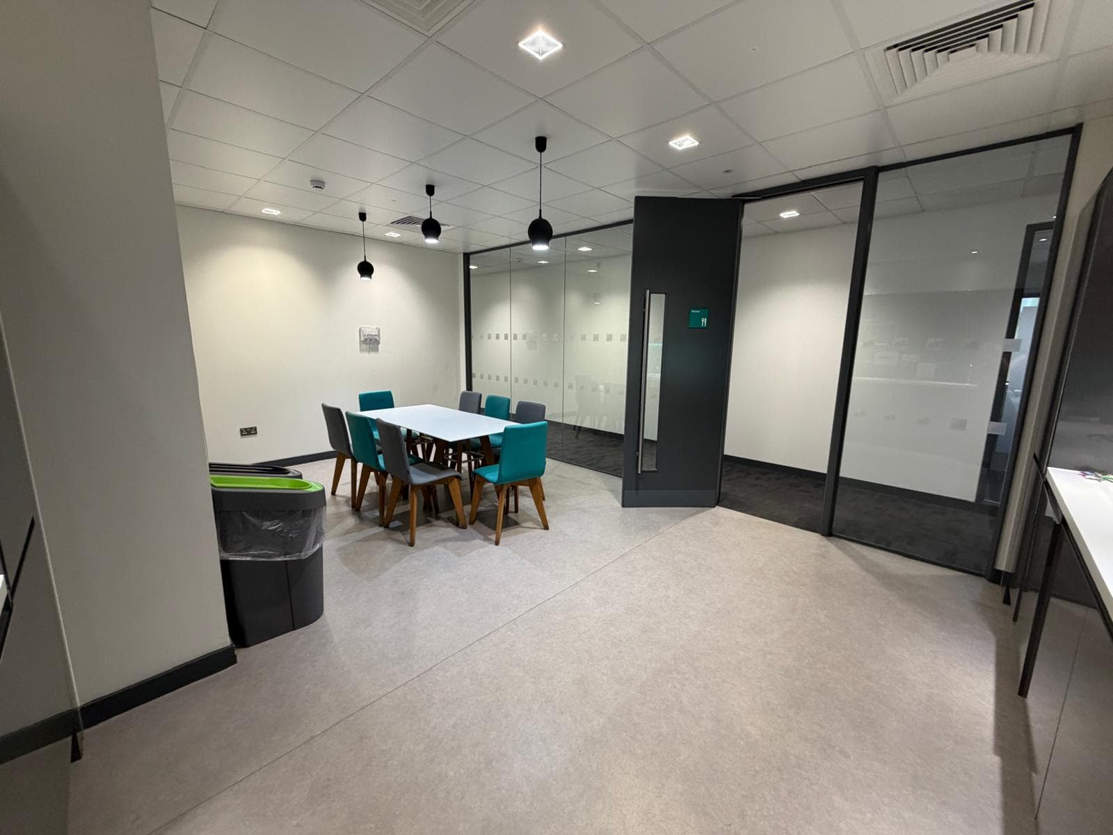 A dining and meeting space with a white table, teal chairs and pendant lights, enclosed by a glazed partition