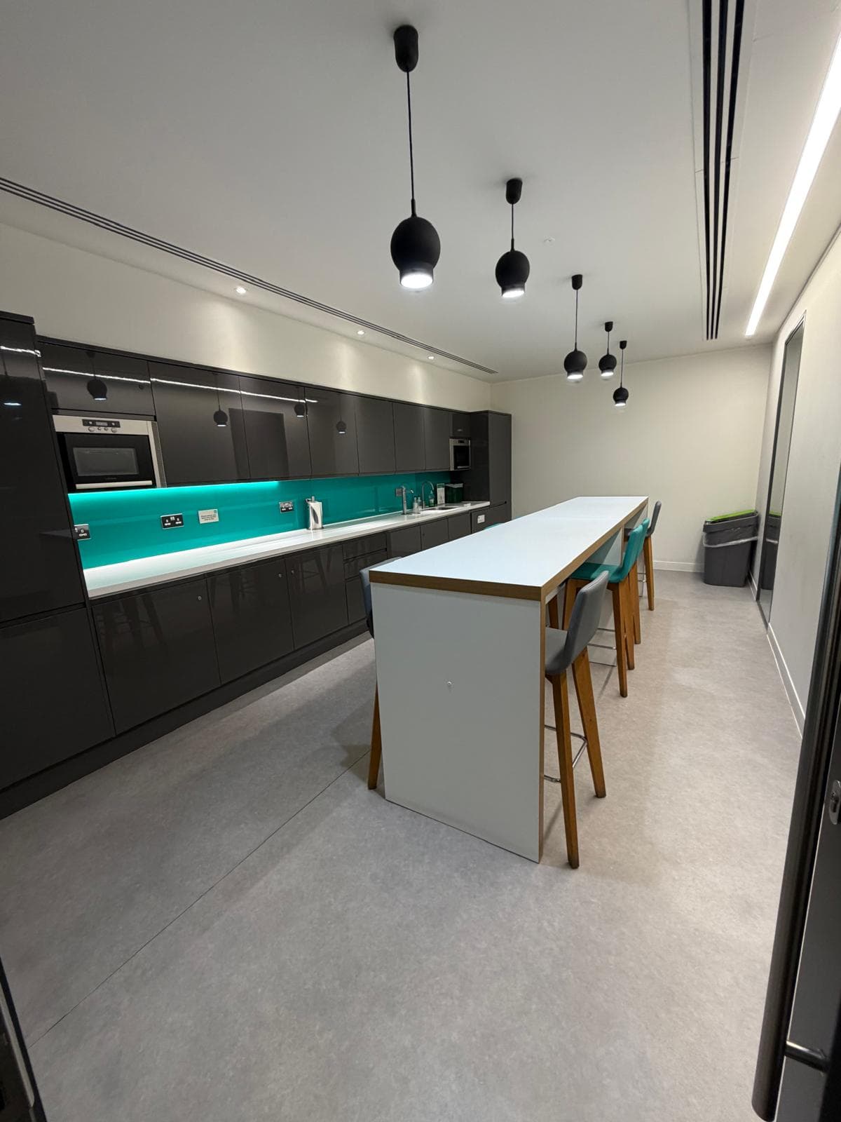 A fitted kitchen with a long run of dark units, integrated oven and lit teal splashback, and a white breakfast bar with teal stools