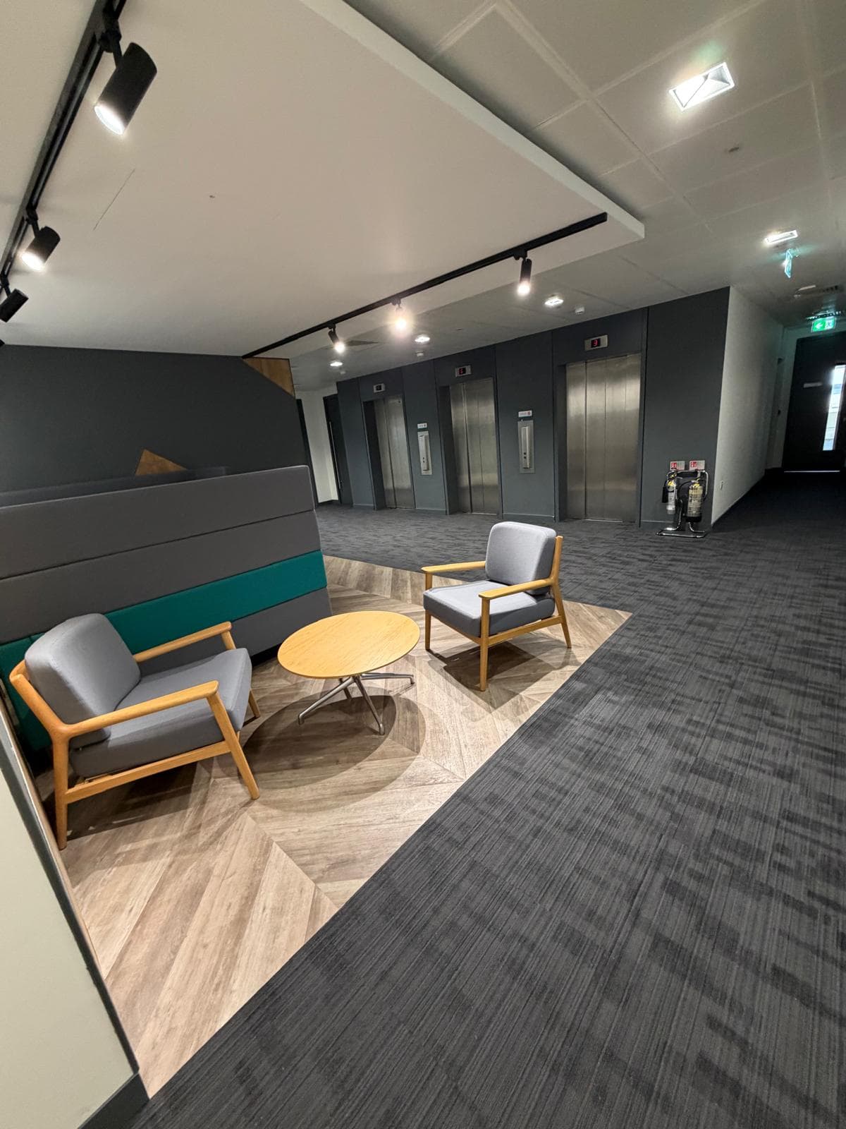 The lift lobby, with steel lift doors, an upholstered feature wall and armchairs around a low table on an inset timber floor