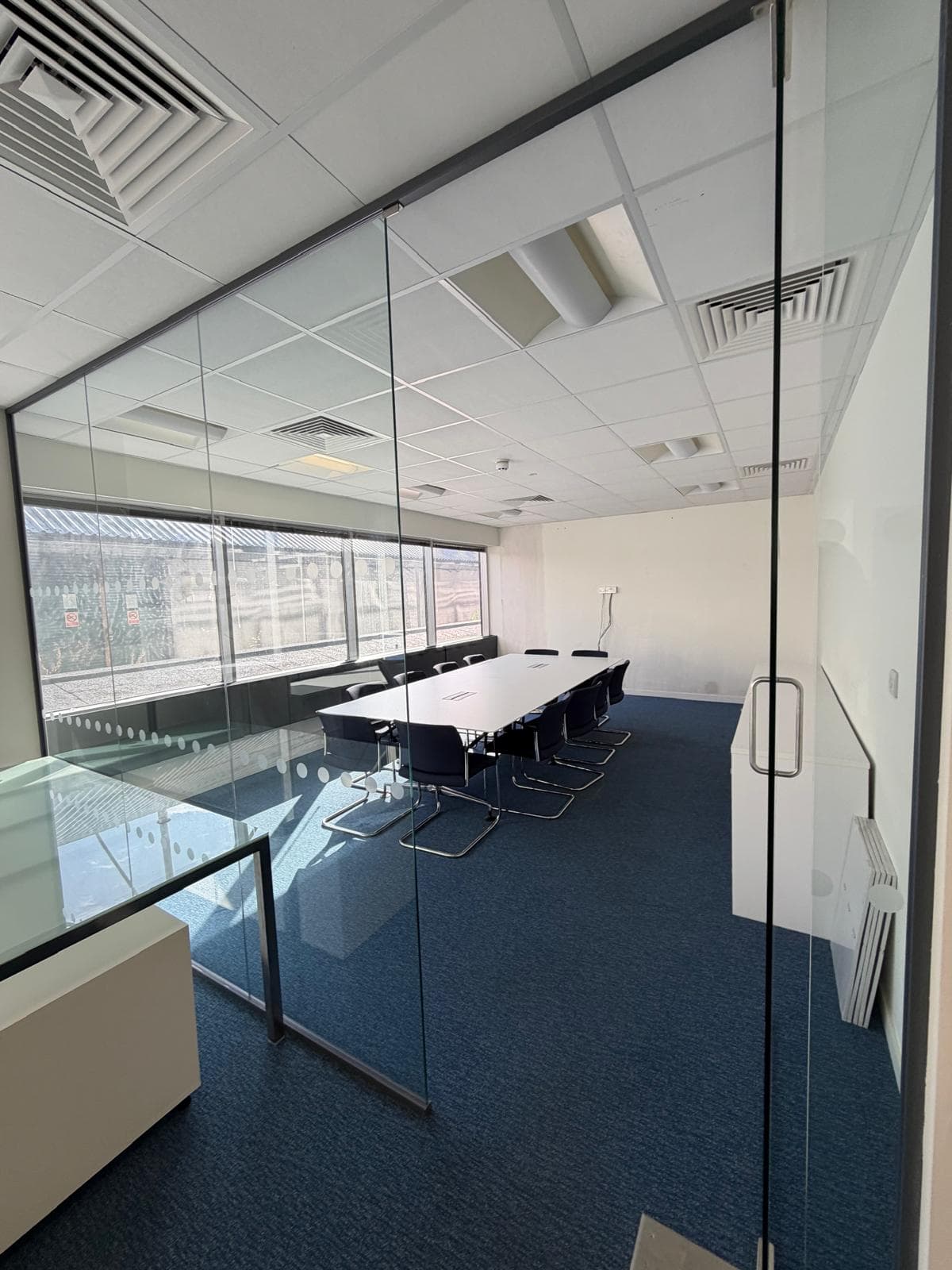A meeting room seen through its glass wall, with a white boardroom table and dark cantilever chairs on navy carpet, lit by a run of windows along one side