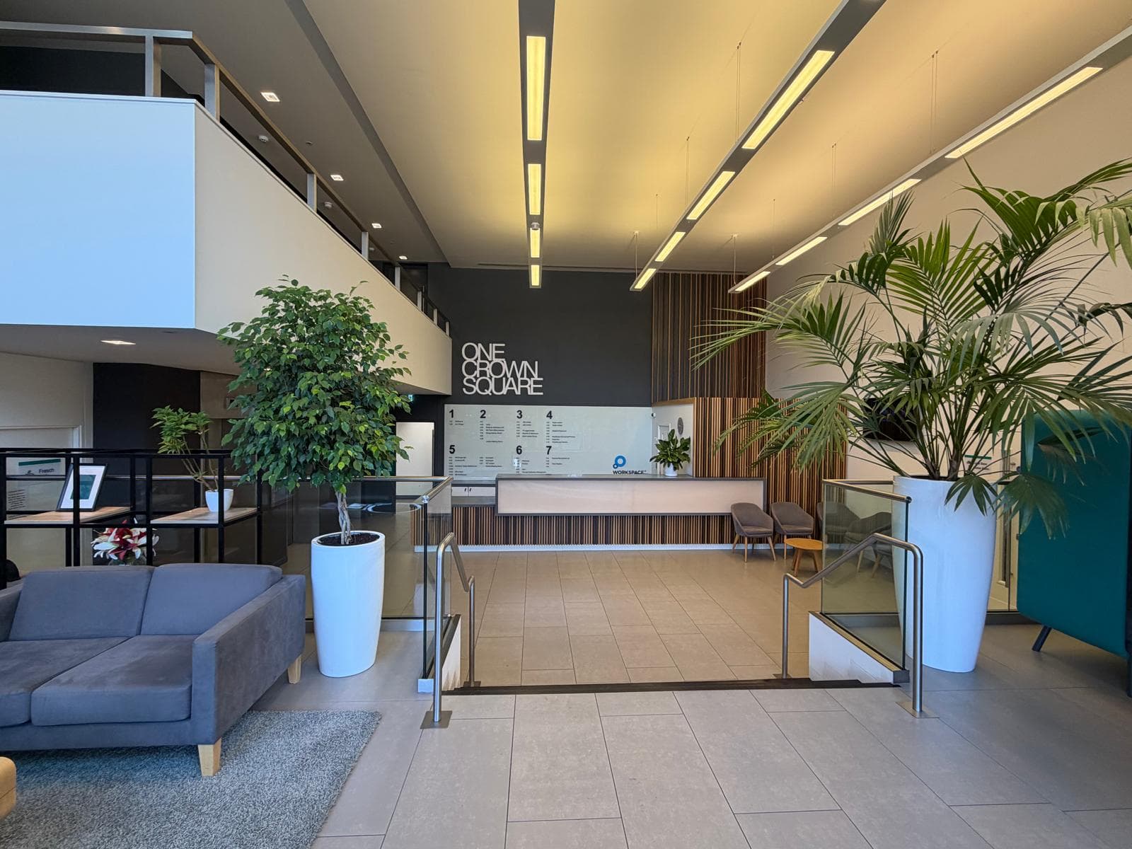 Reception seen past a grey sofa and tall planting, with the mezzanine overhead and the reception desk beyond