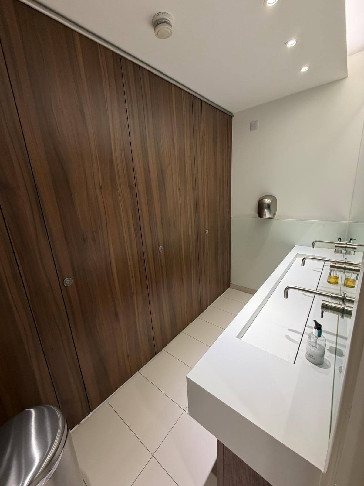 A washroom with full-height walnut cubicle doors and a white solid-surface trough basin