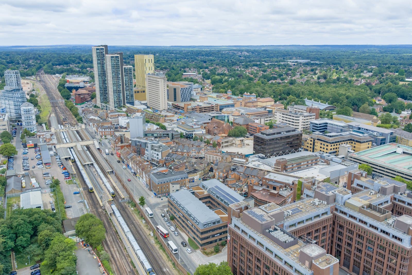 Aerial view over Woking town centre, with the railway station platforms and lines running through the middle of the town and the Surrey countryside beyond